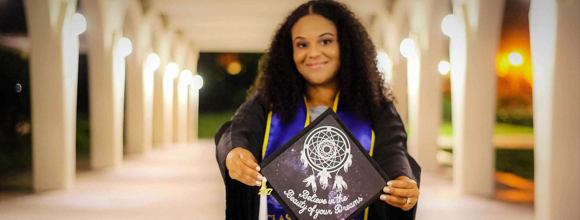 Photo of student holding a graduation cap that says "Believe in the Beauty of Your Dreams."